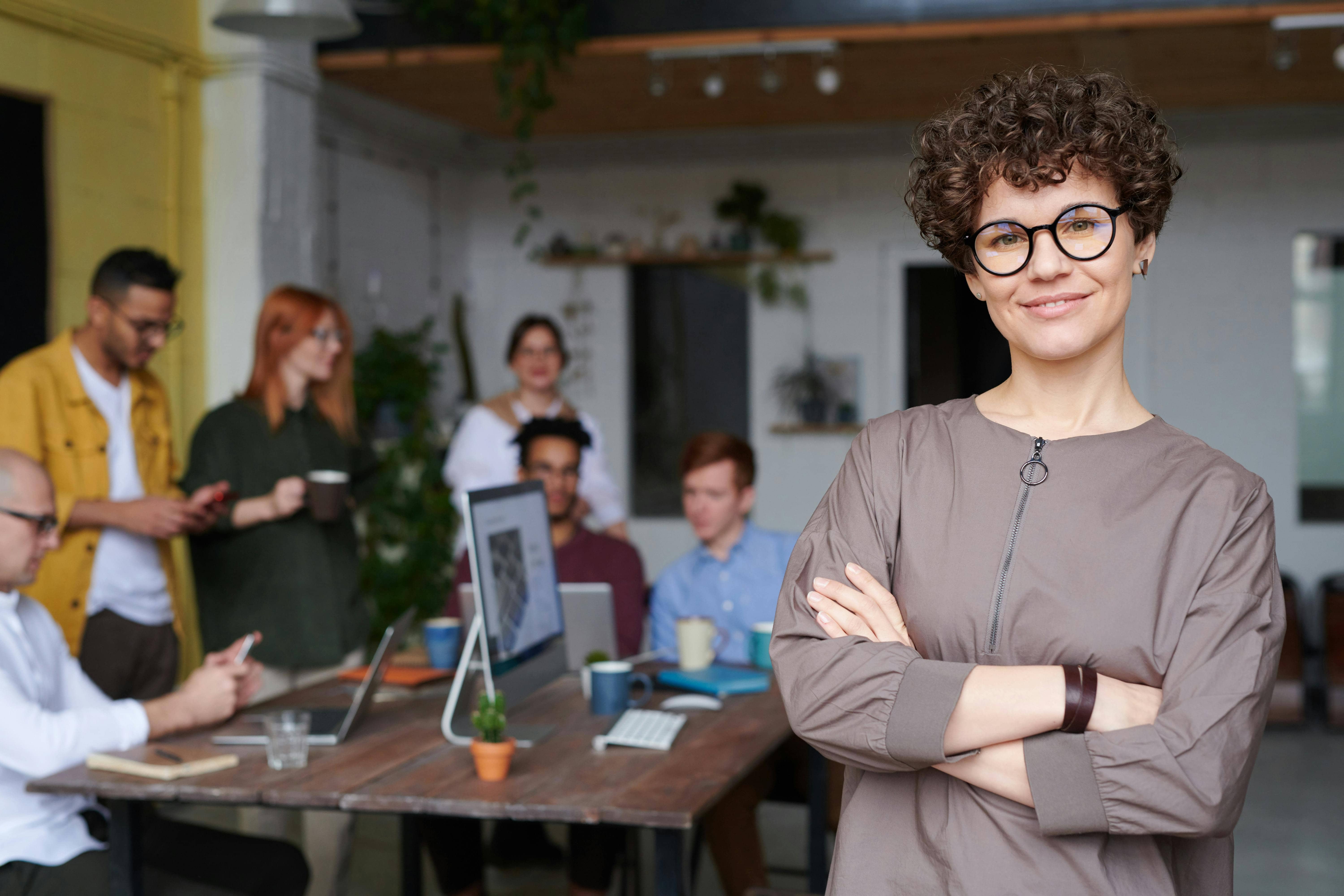 Confident business professional standing with arms crossed in a modern office while a team collaborates in the background