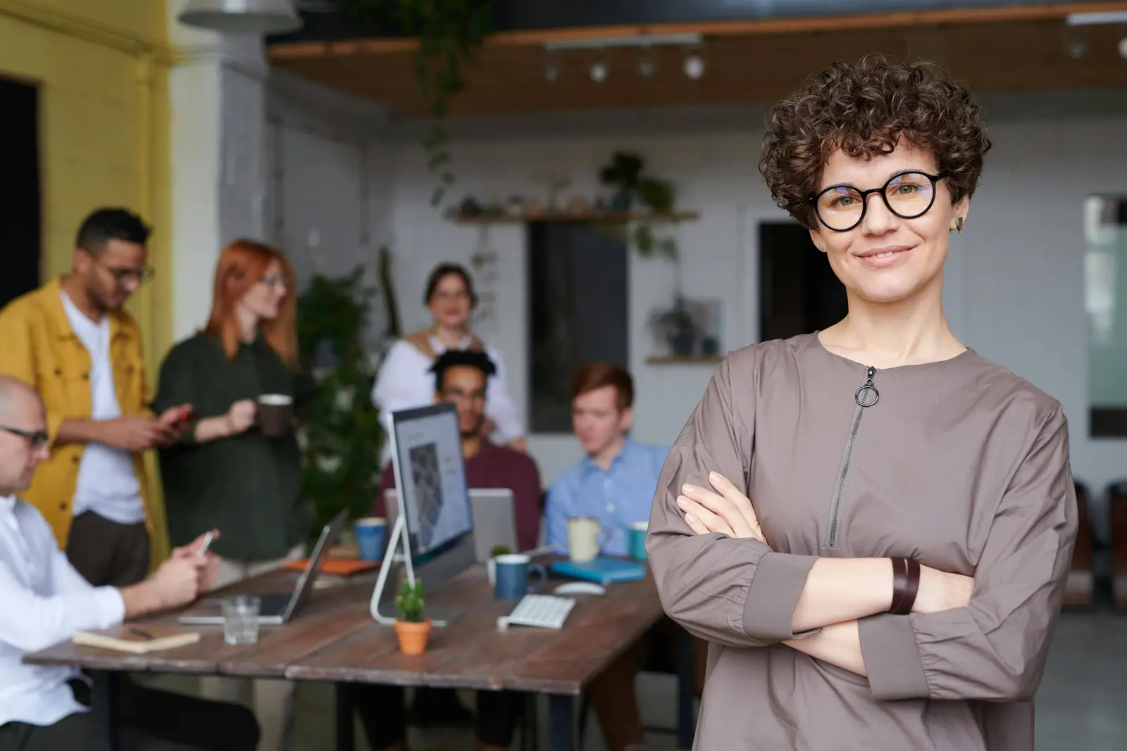 Confident business professional standing with arms crossed in a modern office while a team collaborates in the background
